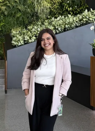 A smiling woman wearing a light pink blazer and white top stands in front of a plant-covered wall.