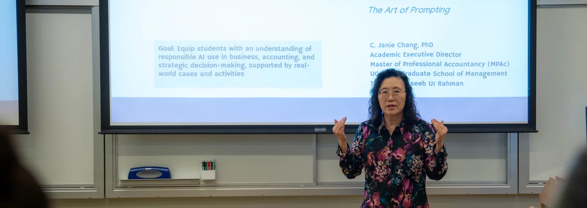 Professor Janie Chang stands in front of a projected presentation titled "AI Transformation" in a classroom, speaking to an audience.