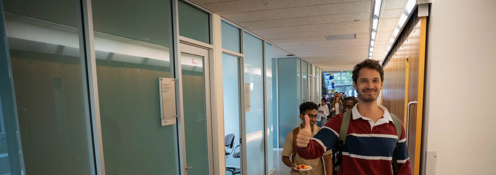 A man in a striped sweater walks down a hallway giving a thumbs up, followed by another person; offices with glass walls line the corridor.