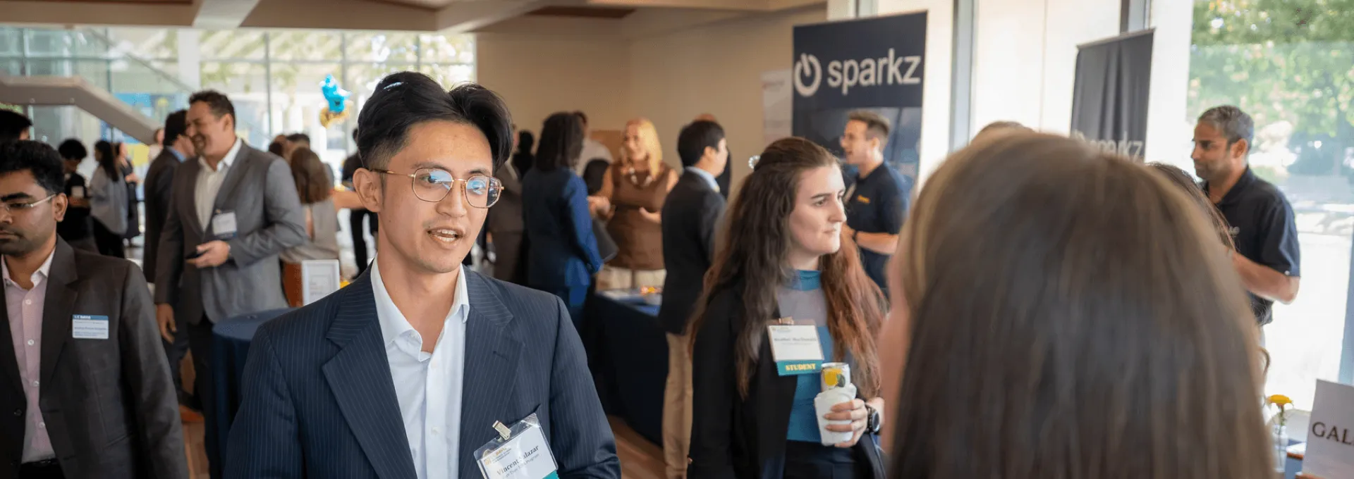 A young man in a suit speaks with a colleague at a crowded professional networking event.