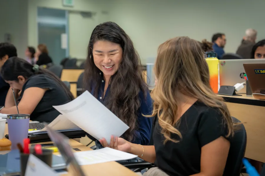 Two women sit in a classroom, smiling and holding papers, with laptops and water bottles on their desks. Other students are working in the background.