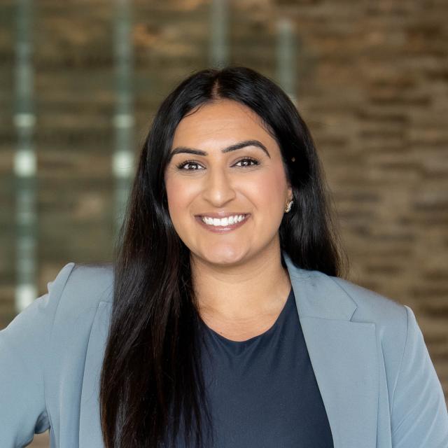 A woman with long dark hair wearing a light gray blazer and dark top stands smiling in front of a blurred brick wall background.