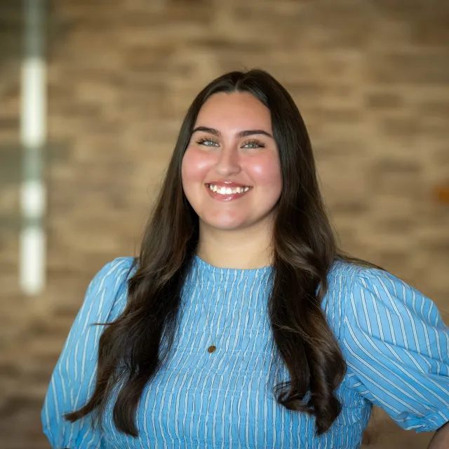 Lizzie Loredo stands smiling in front of a blurred brick wall background.