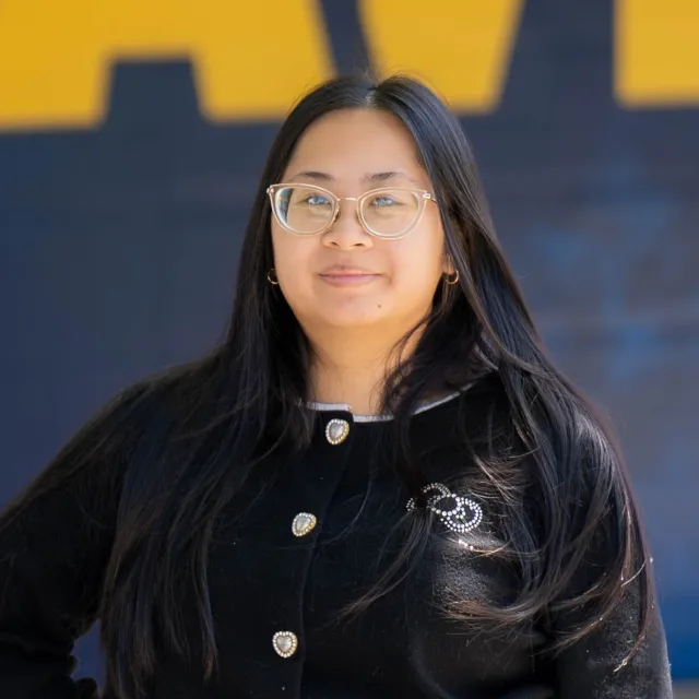 Jenny Nguyen stands in front of a blue and yellow background, wearing a black sweater with decorative buttons.