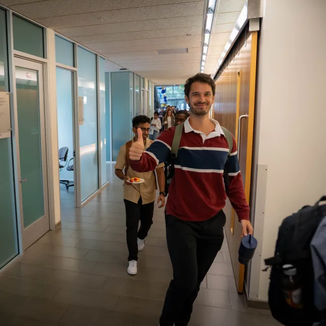 A man in a striped sweater walks down a hallway giving a thumbs up, followed by another person; offices with glass walls line the corridor.