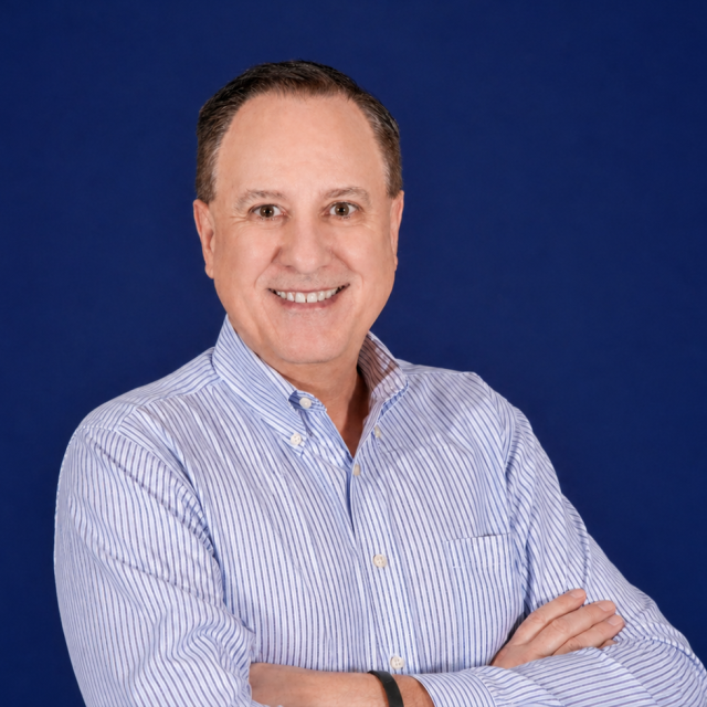 Smiling Tim Akin in a light blue striped shirt stands with arms crossed in front of a plain dark blue background.