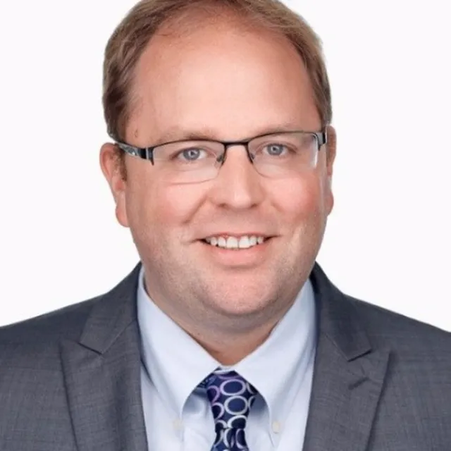 Man with short light brown hair, glasses, and a gray suit jacket over a light blue shirt and patterned tie, smiling at the camera against a plain white background.