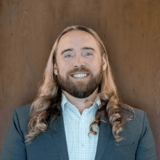 A person with long blond hair and a beard, wearing a suit jacket and checked shirt, stands smiling in front of a wooden background.