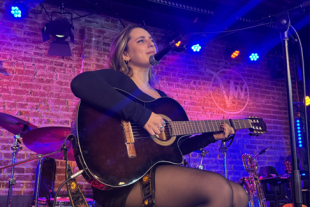 Katya Fomin sitting on a stool on a stage, playing a guitar.