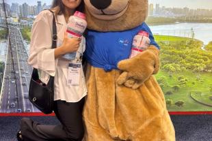 A woman smiles and poses with a person in a bear costume, both holding bottles of Downy Unstopables, in front of a scenic city backdrop.