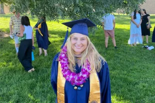 A graduate in a cap and gown with a purple flower lei stands on grass under a tree; other people are gathered in the background at an outdoor event.