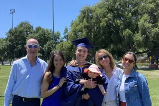 A group of five adults and a baby pose together outdoors on a grassy field. One adult is wearing a graduation cap and gown and holding the baby.