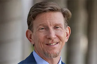 A middle-aged man with light brown hair, a short beard, and mustache is smiling and wearing a blue suit jacket and light blue shirt.