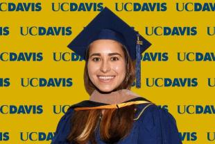 Maria Ahumada wearing a blue graduation cap and gown stands in front of a yellow UC Davis backdrop with repeated logos.