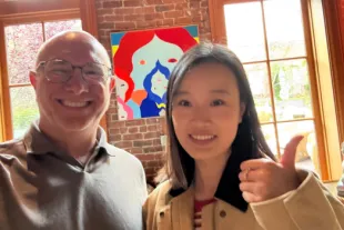 Two people smile for a photo indoors, with a colorful painting and large windows in the background. One person gives a thumbs-up gesture.