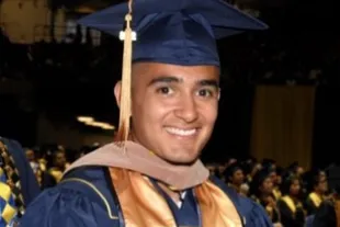 A smiling graduate in a blue cap and gown with a gold stole at a commencement ceremony.