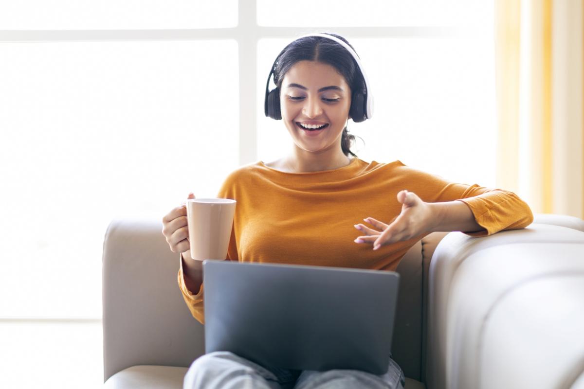 Happy young person making video call via laptop and drinking coffee at home, smiling eastern lady wearing wireless headphones and gesturing at webcamera, sitting on sofa in living room