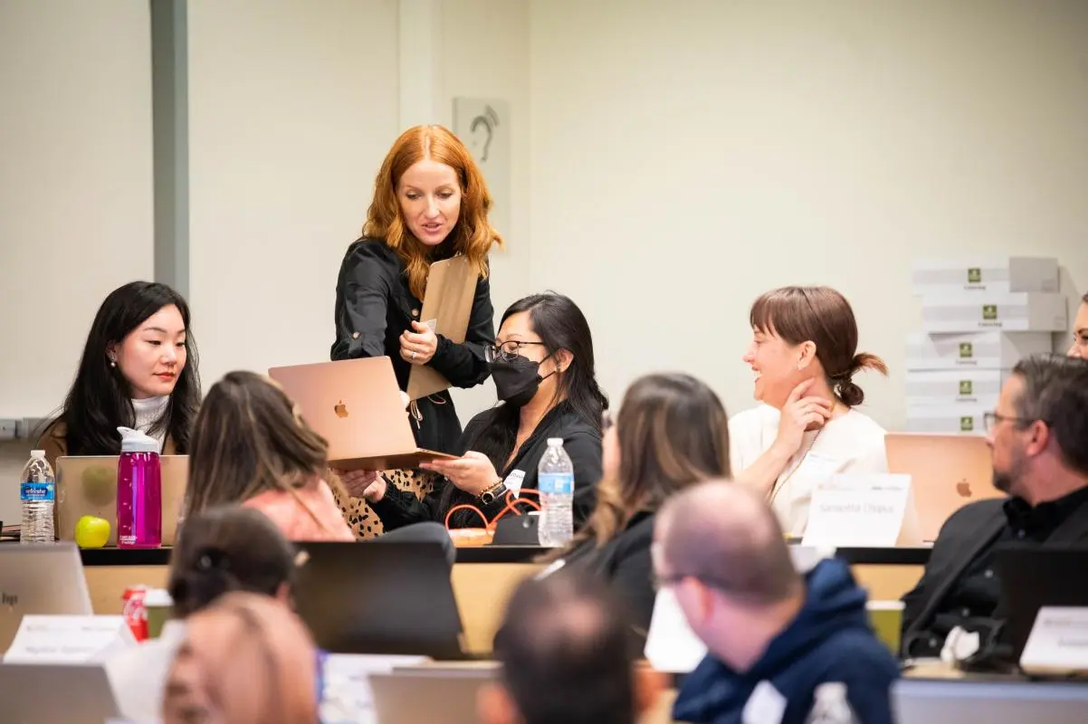 A woman stands and shows her laptop screen to a group of seated people in a classroom setting; others watch and listen attentively.