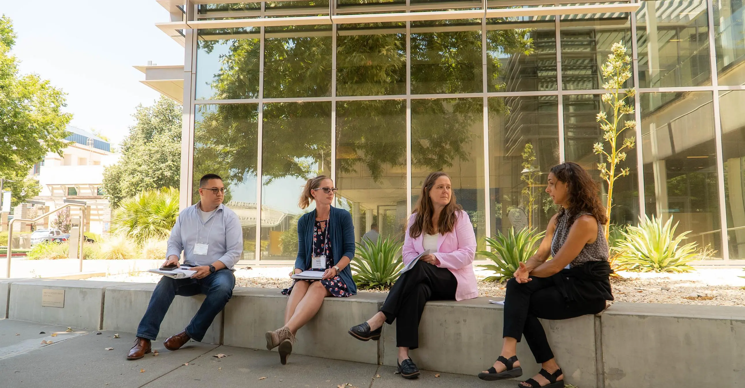 Four people sit on a low concrete wall outside Gallagher Hall, having a discussion with notebooks in hand, on a sunny day.