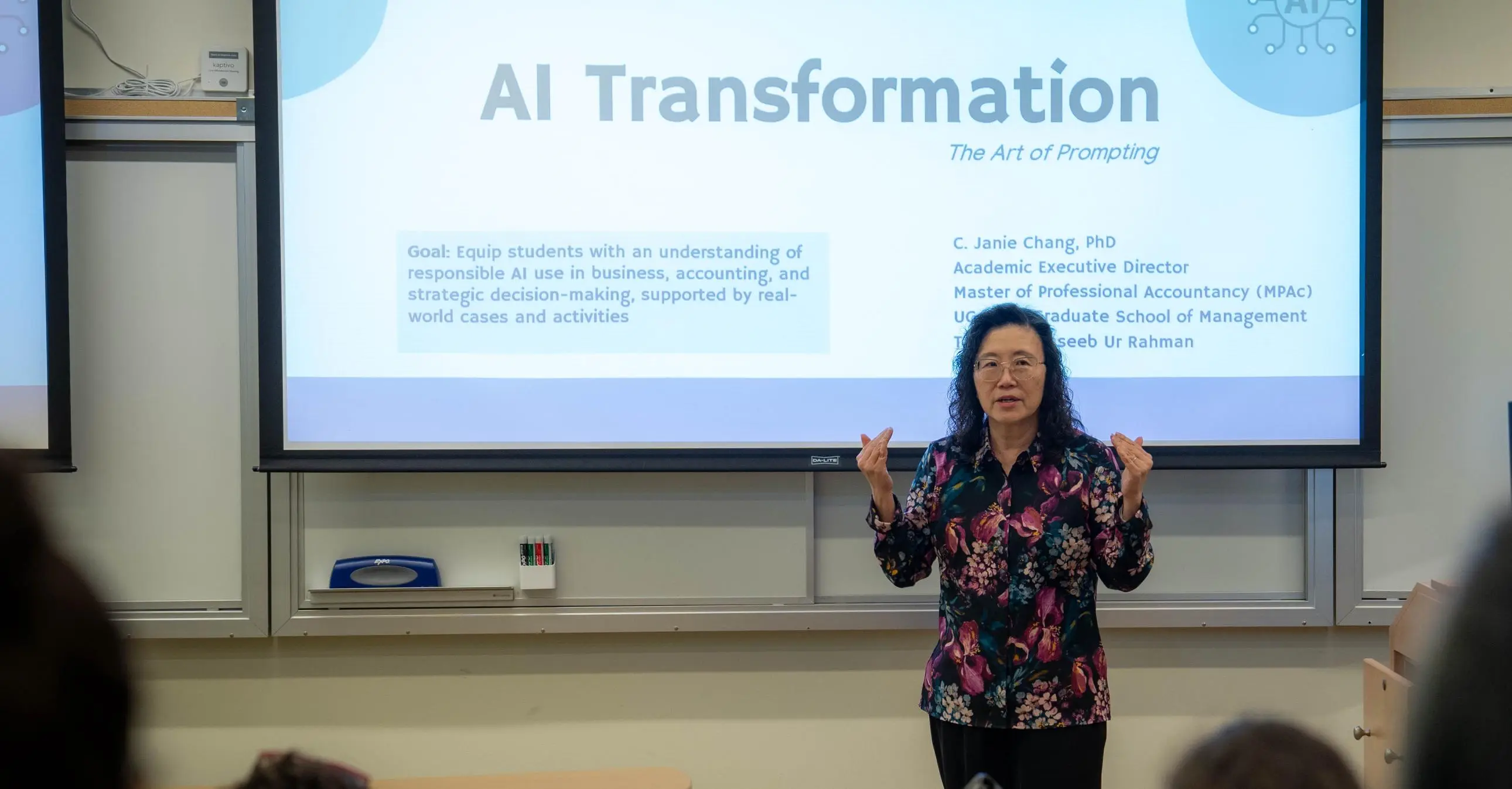 Professor Janie Chang stands in front of a projected presentation titled "AI Transformation" in a classroom, speaking to an audience.