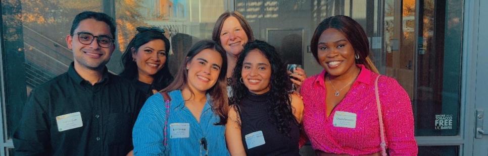 Six people stand together outdoors, smiling at the camera, each wearing name tags. There are drinks and flowers on the table in front of them. Glass windows and sunlight are in the background.