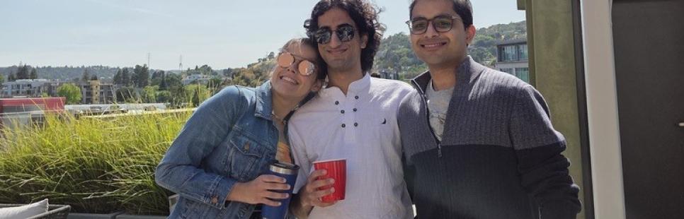 Three people stand together on a sunny rooftop patio, smiling at the camera. Two hold red cups, and outdoor seating is visible in the background.