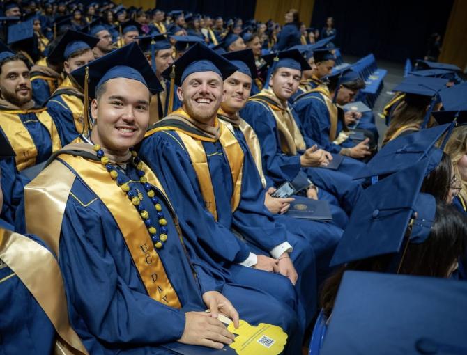 Graduates in blue caps and gowns with gold stoles sit in rows at a commencement ceremony, holding programs and smiling at the camera.