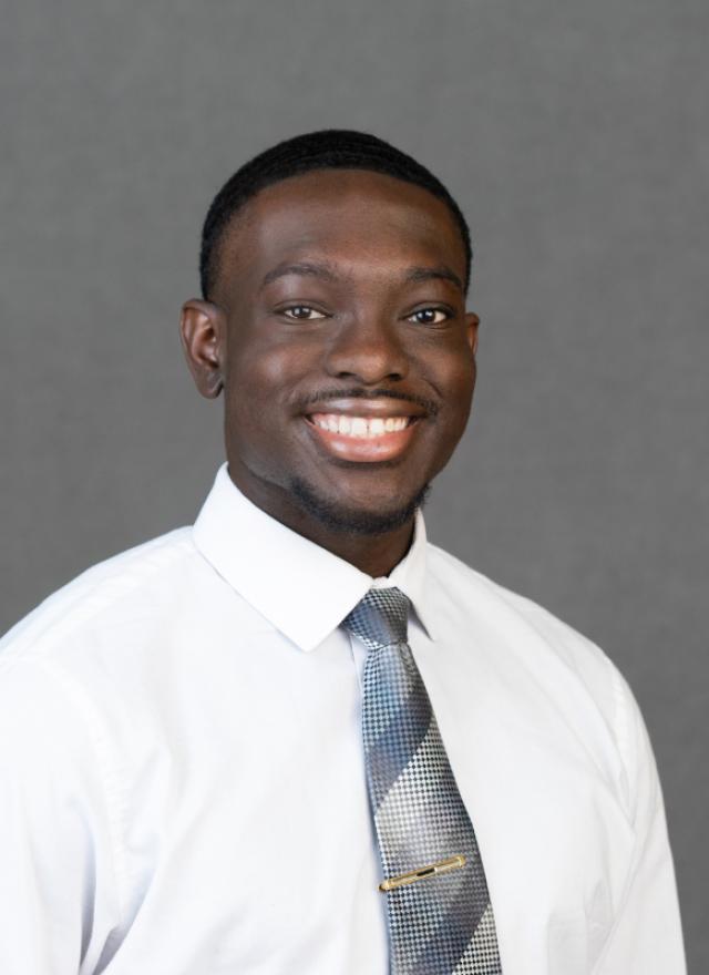 A man wearing a white dress shirt and patterned tie smiles at the camera against a plain gray background.