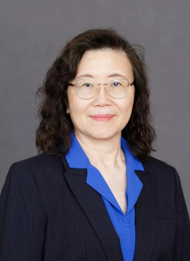 A woman with wavy dark hair, glasses, and a navy pinstripe suit over a blue blouse poses in front of a plain gray background.