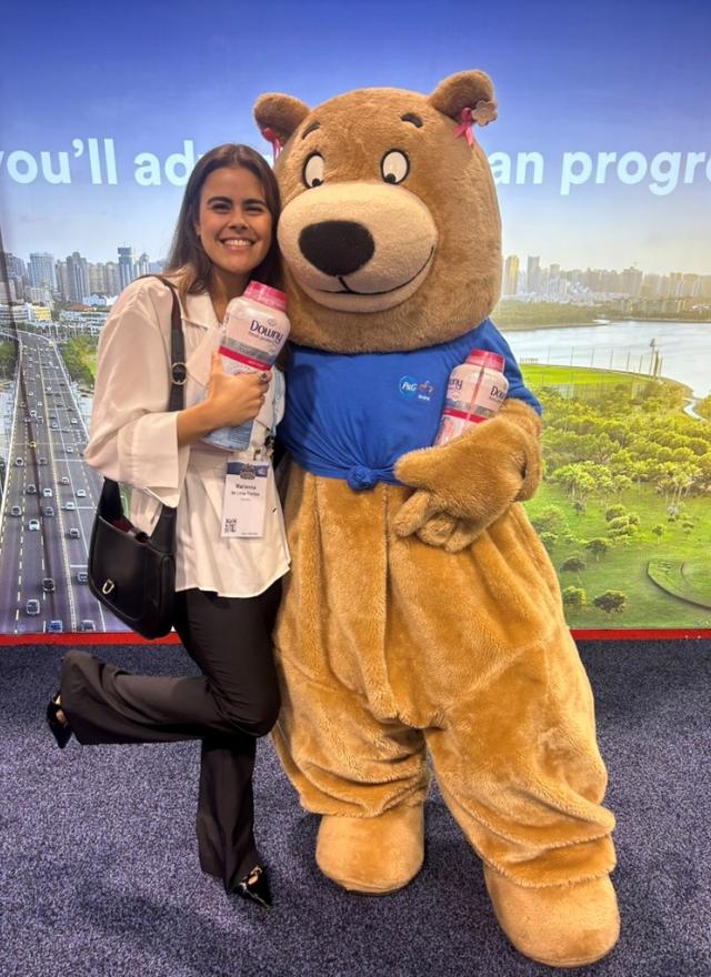 A woman smiles and poses with a person in a bear costume, both holding bottles of Downy Unstopables, in front of a scenic city backdrop.