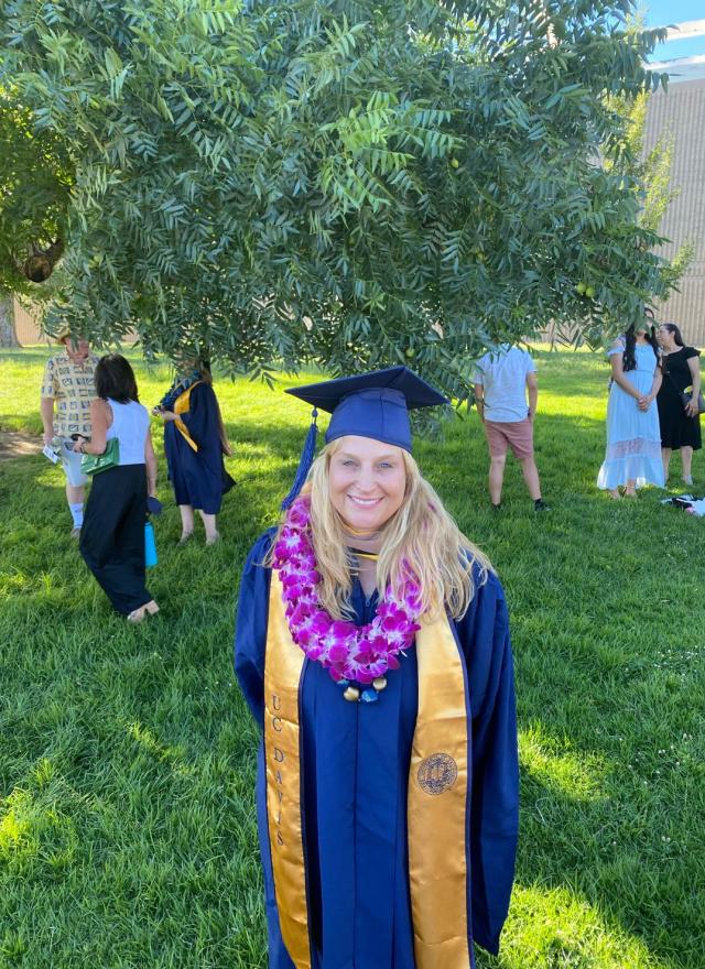A graduate in a cap and gown with a purple flower lei stands on grass under a tree; other people are gathered in the background at an outdoor event.