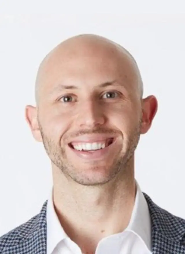 A smiling bald man wearing a white shirt and a checkered blazer, posed against a plain white background.