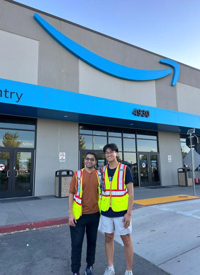 Two people wearing safety vests stand in front of an Amazon building entrance with the Amazon logo and address 4930 visible above them.