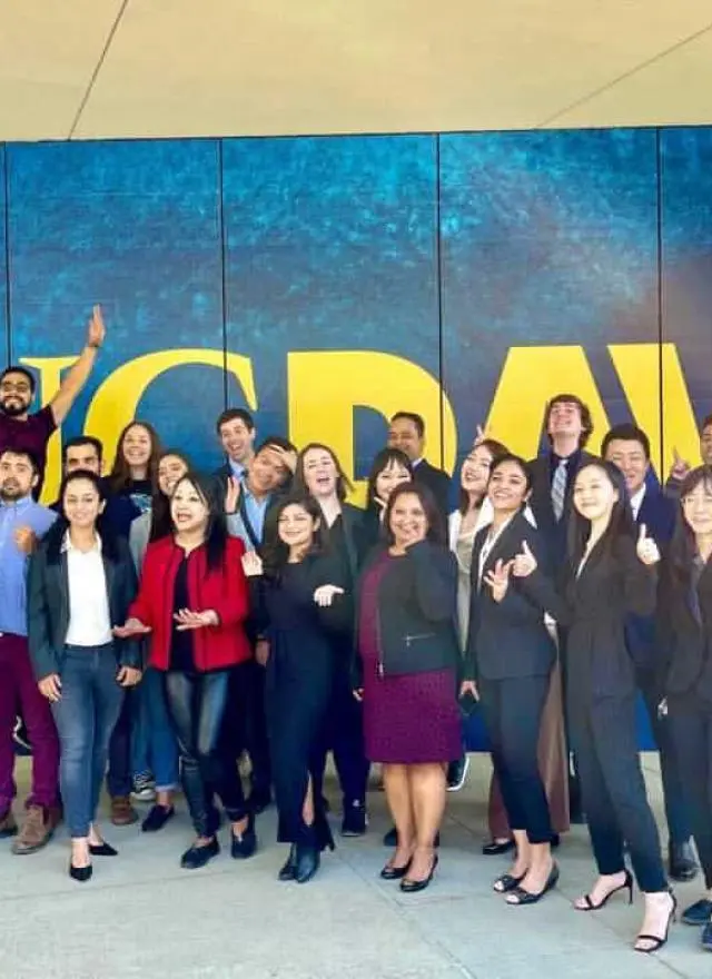A group of people posing and smiling in front of a large blue and yellow “UC DAVIS” sign, with several making enthusiastic gestures.