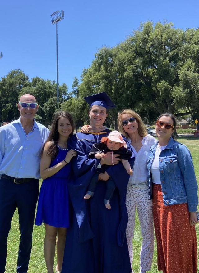 A group of five adults and a baby pose together outdoors on a grassy field. One adult is wearing a graduation cap and gown and holding the baby.