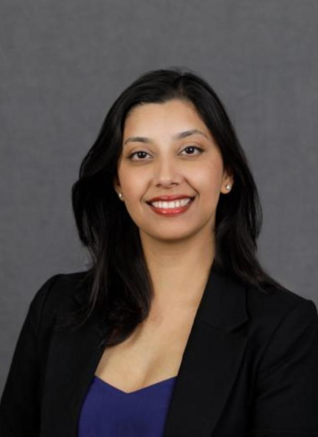 A woman with long dark hair wearing a black blazer and blue top smiles in front of a plain gray background.