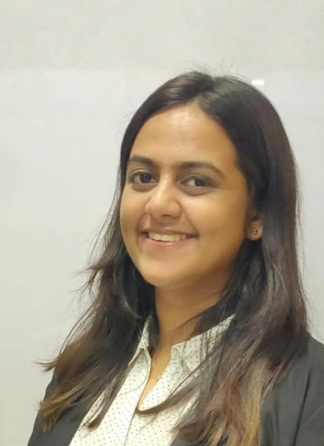 A woman with long dark hair, wearing a white blouse and black blazer, smiles while standing in front of a plain light-colored background.