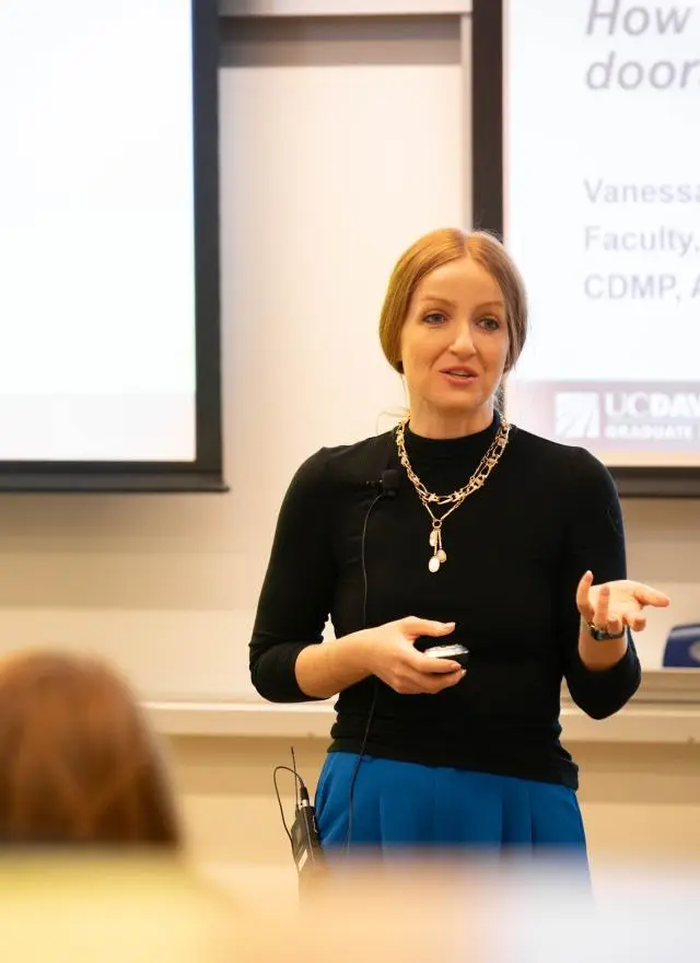 A woman stands in front of a classroom giving a presentation, gesturing with one hand. A projection screen and audience members are visible.