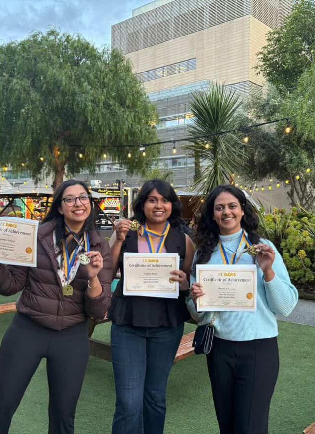 Three women stand outdoors holding certificates and wearing medals, smiling at the camera. There are picnic tables, plants, and a building in the background.
