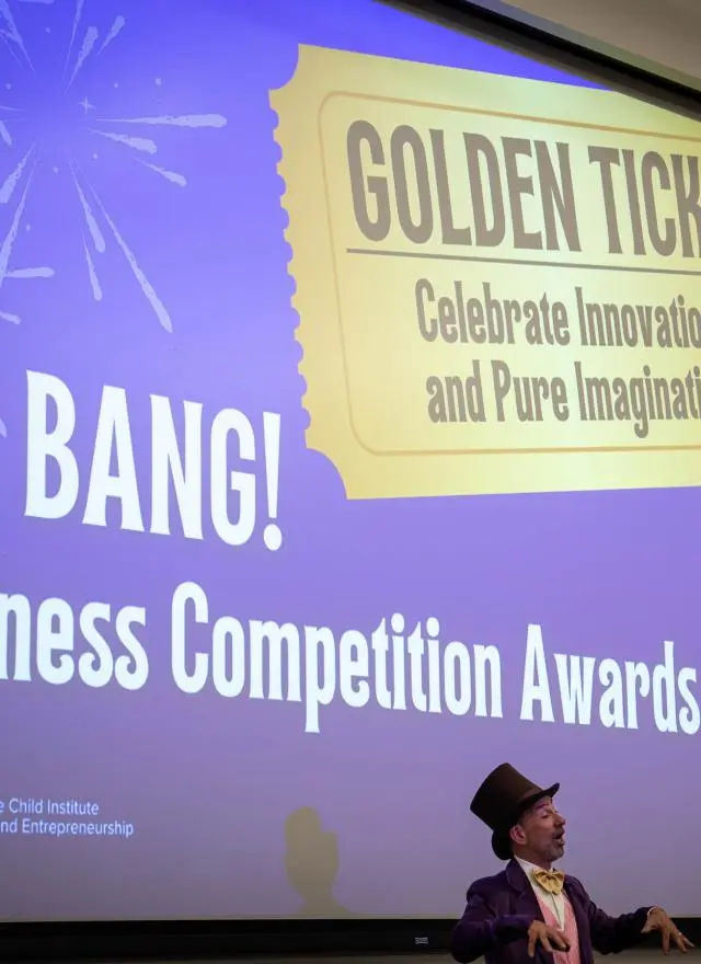 A person in a Willy Wonka costume stands in front of a projection reading "BIG BANG! Business Competition Awards" and "GOLDEN TICKET Celebrate Innovation and Pure Imagination.