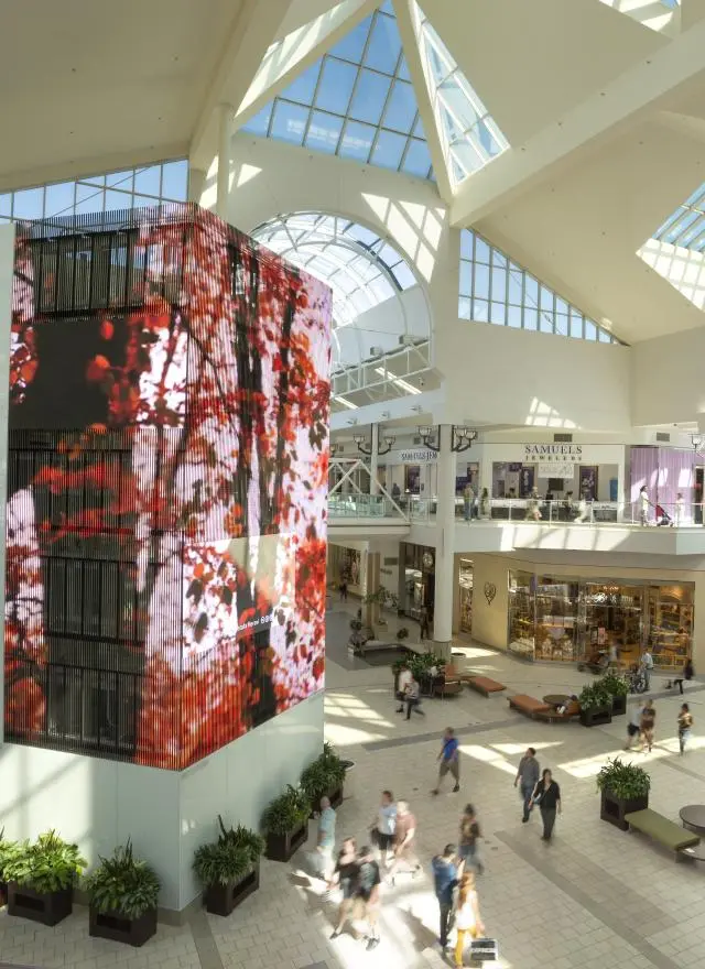 Spacious, modern shopping mall interior with a glass ceiling, digital display, multiple stores, and people walking on two levels.