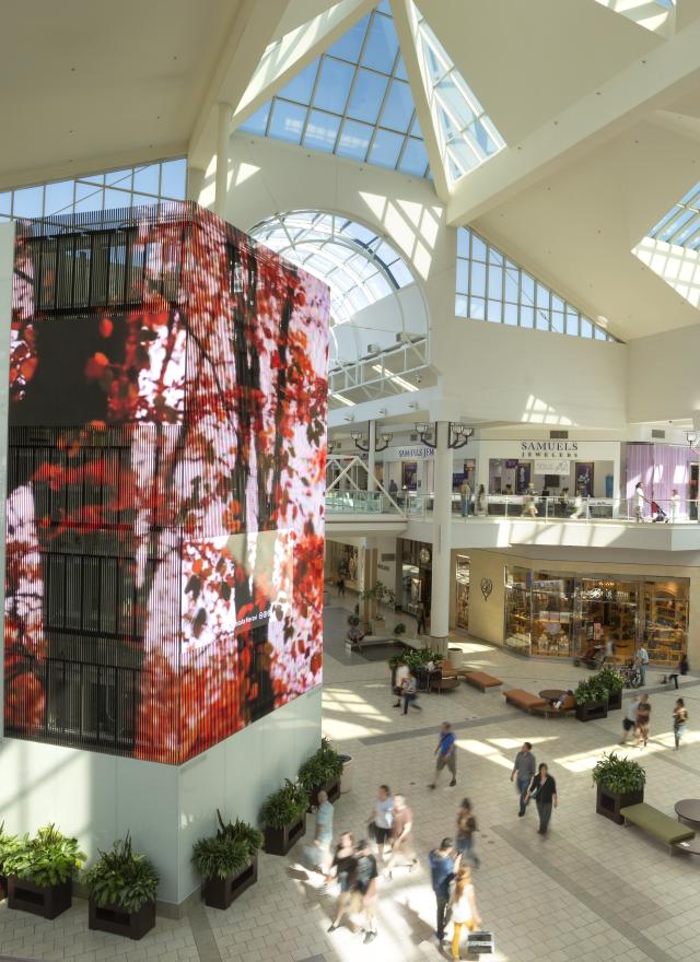 Spacious, modern shopping mall interior with a glass ceiling, digital display, multiple stores, and people walking on two levels.