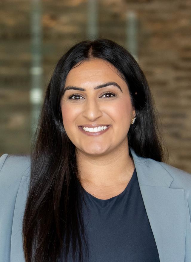 A woman with long dark hair wearing a light gray blazer and dark top stands smiling in front of a blurred brick wall background.