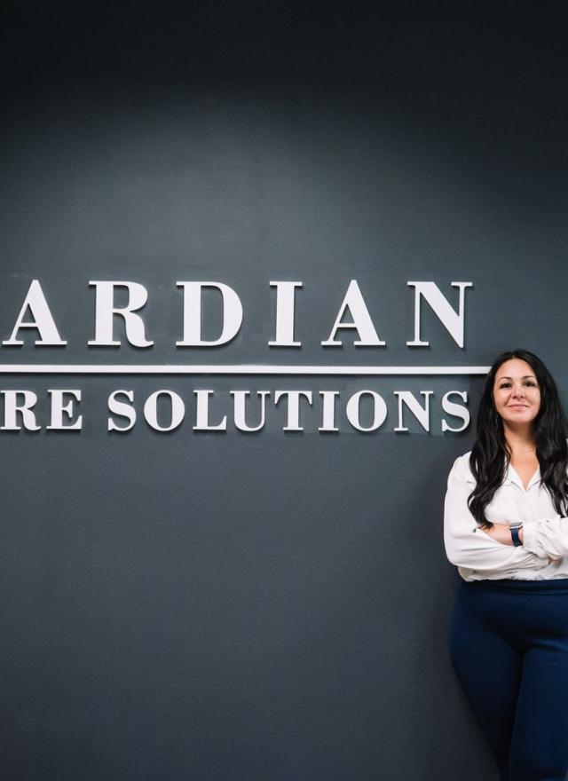 A woman stands with arms crossed in front of a dark wall displaying the words "Guardian Secure Solutions.