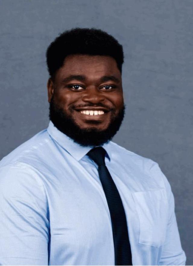 A man with a beard and afro hairstyle, wearing a light blue dress shirt and dark tie, smiles in front of a plain gray background.