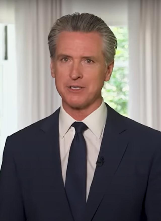 Gavin Newsom in a navy suit and tie stands indoors between two windows, with American and California state flags visible in the background.