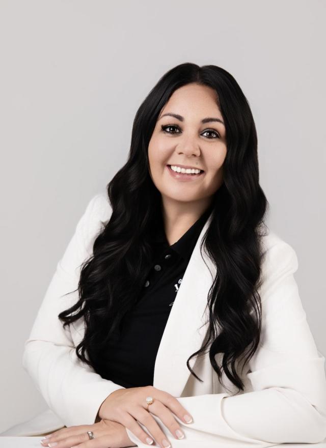 A woman with long dark hair, wearing a white blazer over a black top, is seated and smiling at the camera against a plain light background.