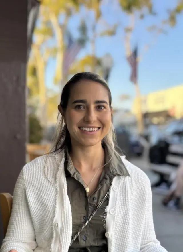 A woman with long hair, wearing a white sweater over a green shirt, sits outdoors at a restaurant table, smiling at the camera. Trees and parked cars are visible in the background.