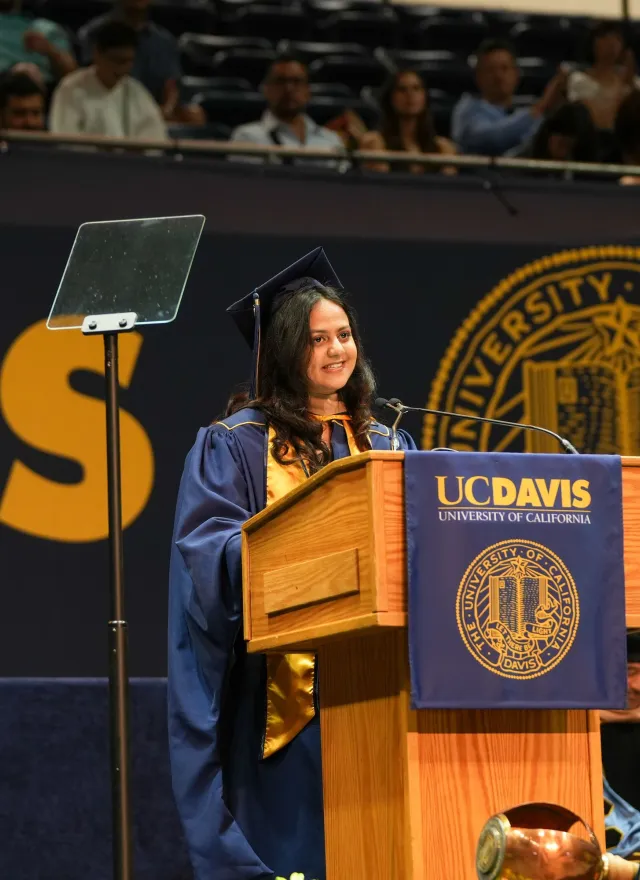 A student in cap and gown speaks at a podium during a UC Davis graduation ceremony, with faculty seated behind and audience in the background.