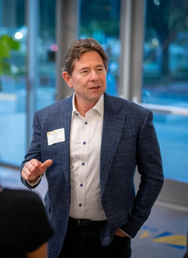 A man in a suit jacket talks to three people at an indoor event, wearing a name tag, with large windows and plants in the background.